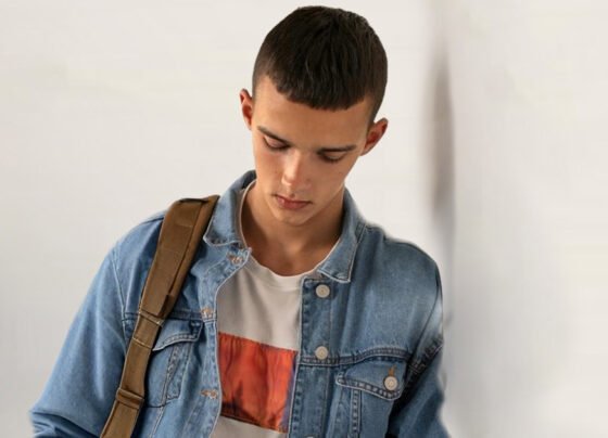 Um adolescente com o ombro encostado em uma parede e uma bolsa pendurada olha para baixo. Tem cabelo bem curto e usa jaqueta jeans