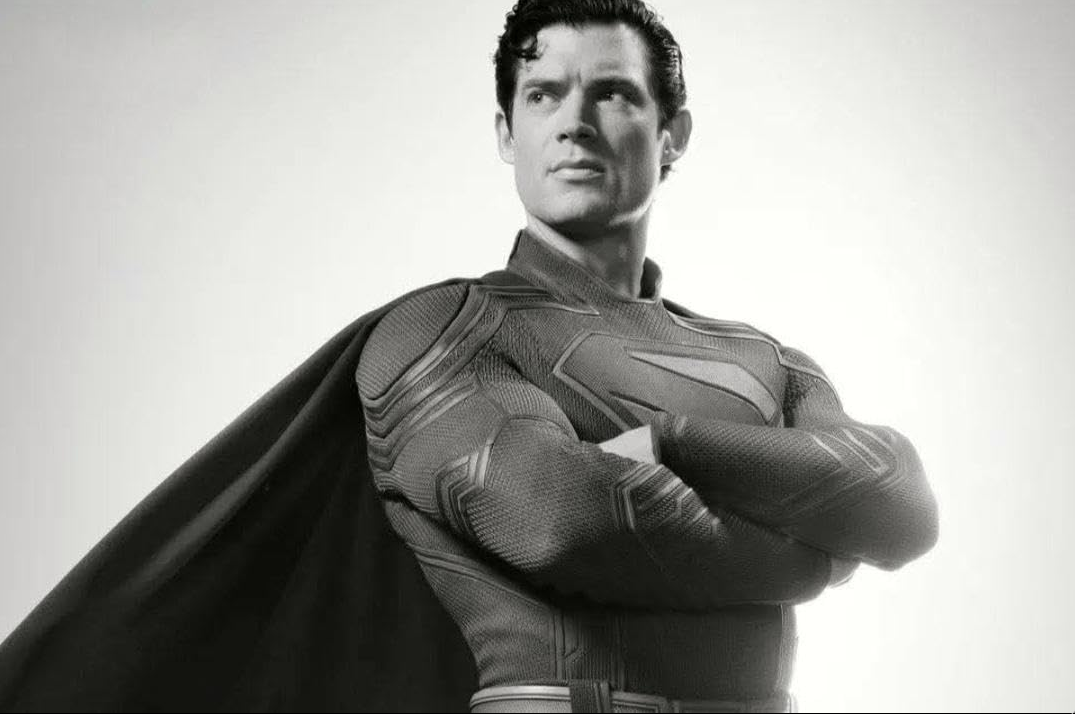 A imagem é um retrato em preto e branco de um homem vestido como Super -Homem. Ele está de pé com os braços cruzados na frente dele e está olhando diretamente para a câmera com uma expressão séria no rosto. O homem está usando seu icônico terno vermelho e azul com o logotipo do Super -Homem no peito. Sua capa está traçando atrás dele, cobrindo o ombro esquerdo. O fundo é claro e branco, tornando o homem o ponto focal da imagem.