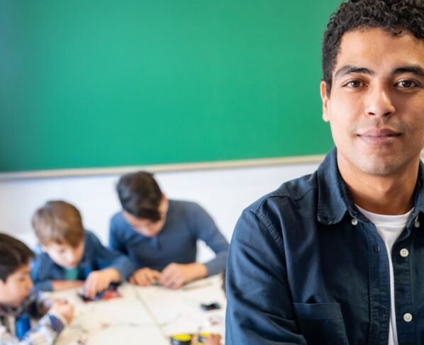 Professor jovem em destaque, de braços cruzados, olhando diretamente para a câmera em uma sala de aula. Ao fundo, um grupo de crianças está sentado em uma mesa, concentrado em uma atividade manual. Ao fundo há um grande quadro verde e paredes com cartazes coloridos parcialmente visíveis. O foco está no educador, transmitindo segurança e protagonismo.