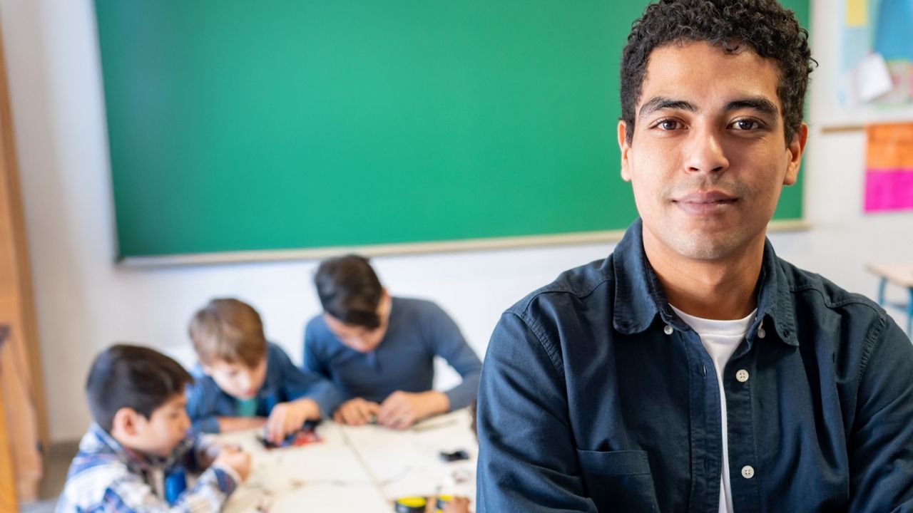 Professor jovem em destaque, de braços cruzados, olhando diretamente para a câmera em uma sala de aula. Ao fundo, um grupo de crianças está sentado em uma mesa, concentrado em uma atividade manual. Ao fundo há um grande quadro verde e paredes com cartazes coloridos parcialmente visíveis. O foco está no educador, transmitindo segurança e protagonismo.